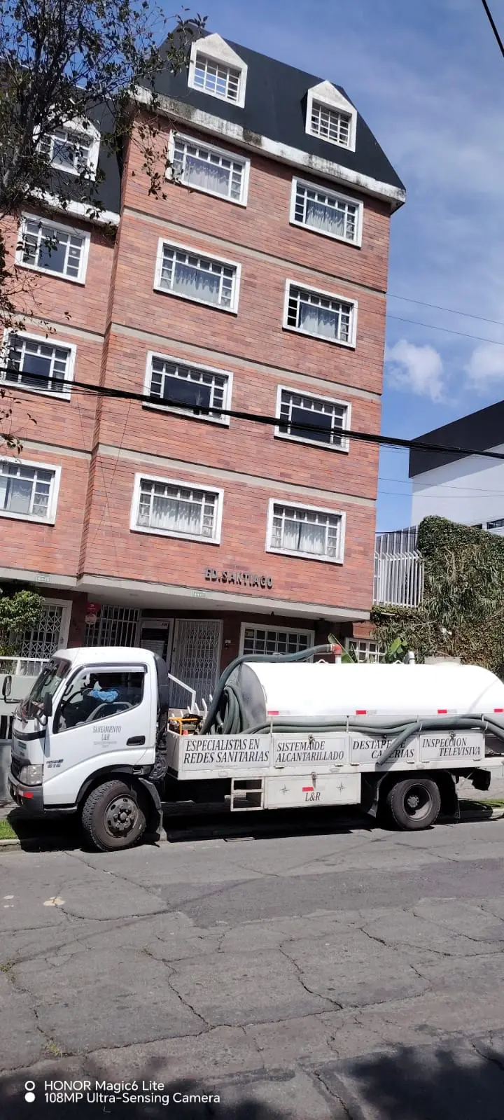 Camión cisterna de Saneamiento RAMGL estacionado frente a un edificio residencial de ladrillo para realizar mantenimiento de alcantarillado.