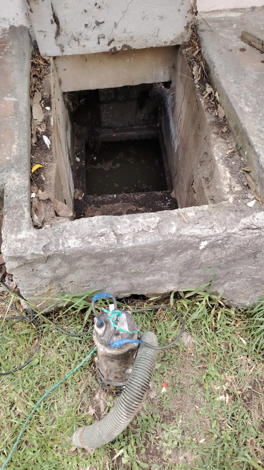 Bomba sumergible portátil instalada sobre el césped junto a la abertura abierta de una cisterna subterránea de agua potable para su vaciado y posterior limpieza.