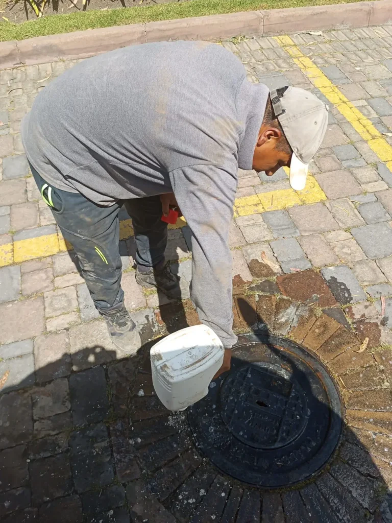 Hombre vertiendo líquido desde un bidón blanco en una alcantarilla circular sobre un suelo de adoquines.